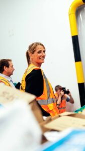 Visitor wearing a high-visibility vest touring a food redistribution facility, with equipment and donated goods visible in the foreground.